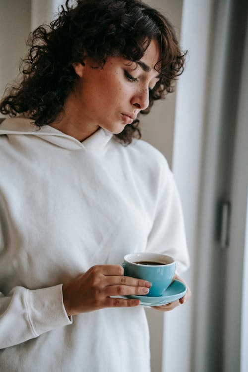 Free Disappointed woman with curly hair standing with ceramic mug of hot drink and looking down Stock Photo