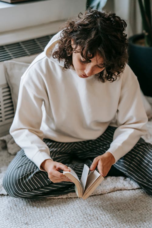 High angle of focused female in casual outfit flipping pages of book while sitting on floor at home