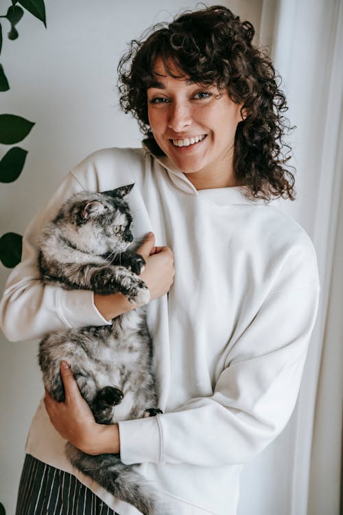 Positive female with curly hair wearing white hoodie cuddling short haired cat and looking at camera