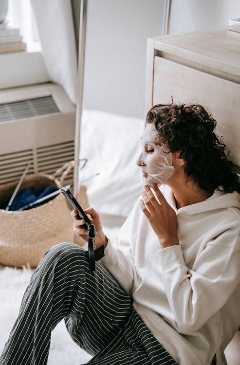 High angle of female surfing Internet while doing beauty procedure with moisturizing sheet mask at home