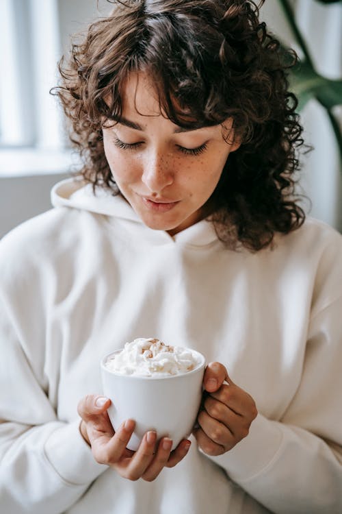 Young female with curly hair and freckles blowing on whipped cream of hot beverage