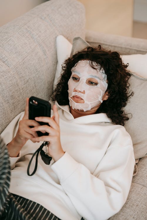 From above of female in moisturizing beauty mask applied on face lying on couch and browsing cellphone