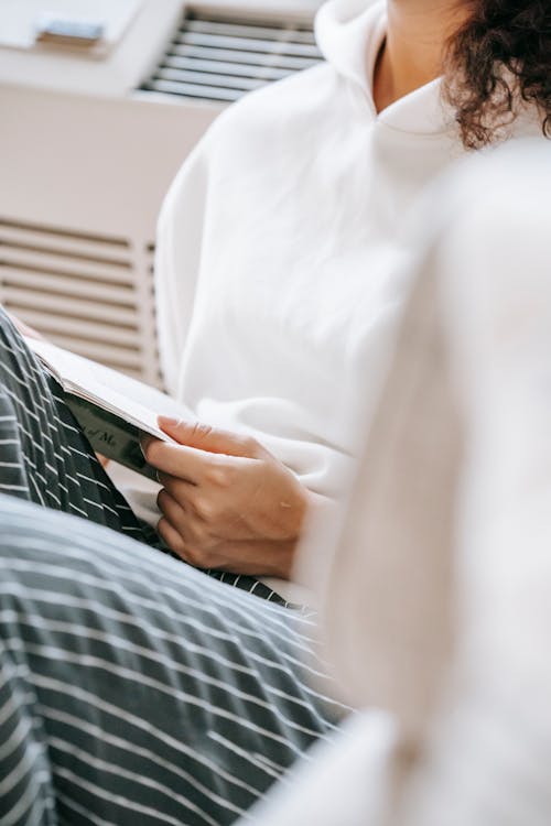 Free Unrecognizable female with dark hair in white sweater and striped pants reading novel while sitting in room during weekend at home Stock Photo