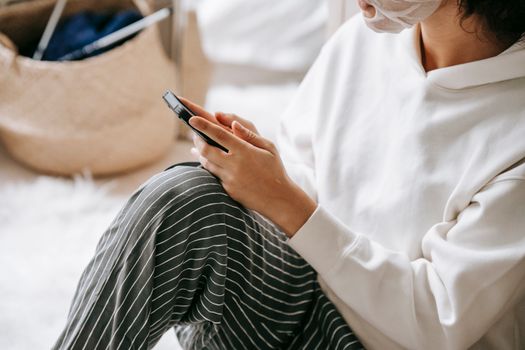 From above of unrecognizable female in sheet mask surfing cellphone while sitting on floor with bent leg in light room on blurred background