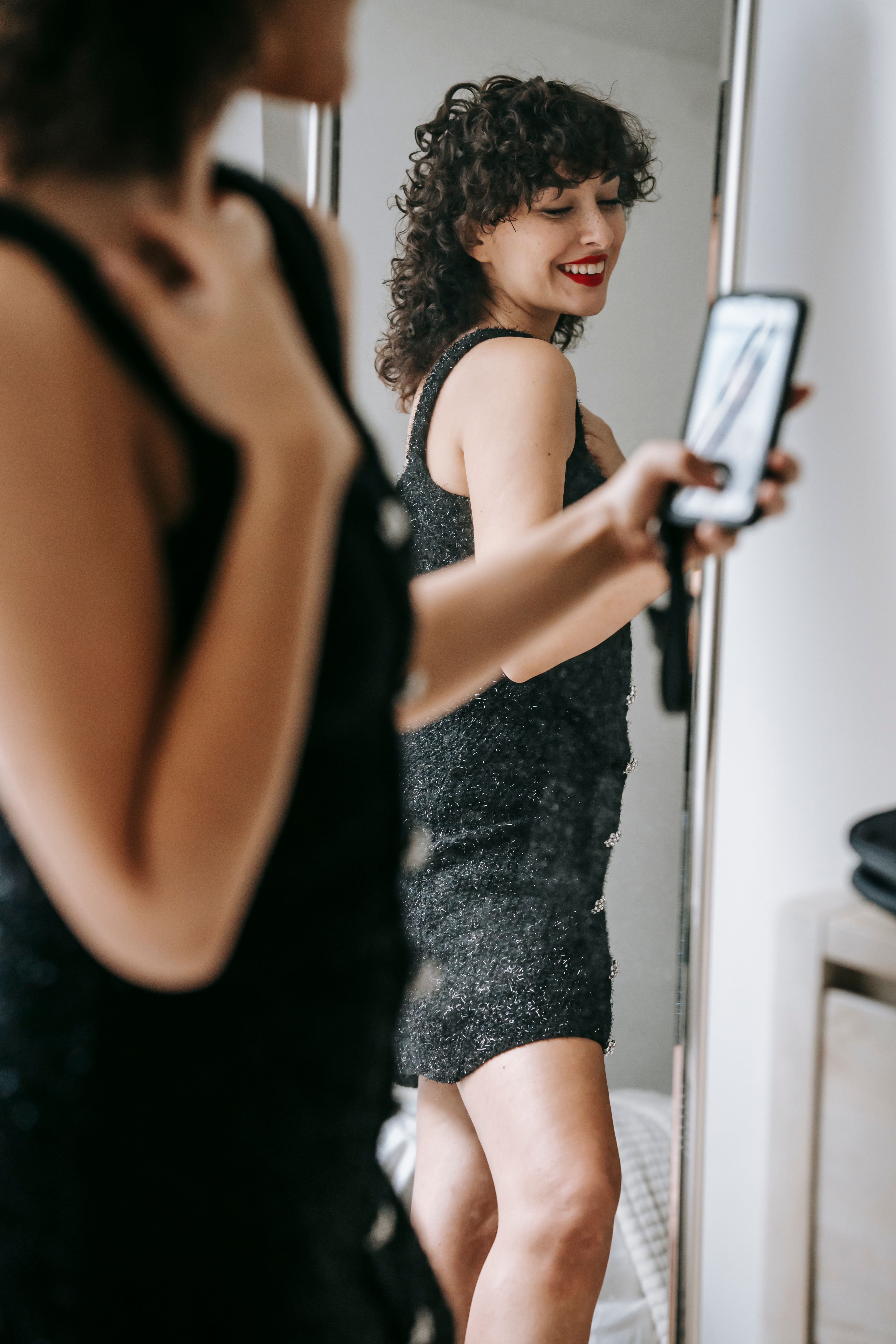 Side view of smiling female with red lips in black dress standing against mirror while taking self p