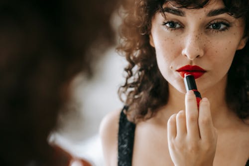 Attractive female with freckles and dark curly hair looking at mirror while applying red lipstick during cosmetic routine in room
