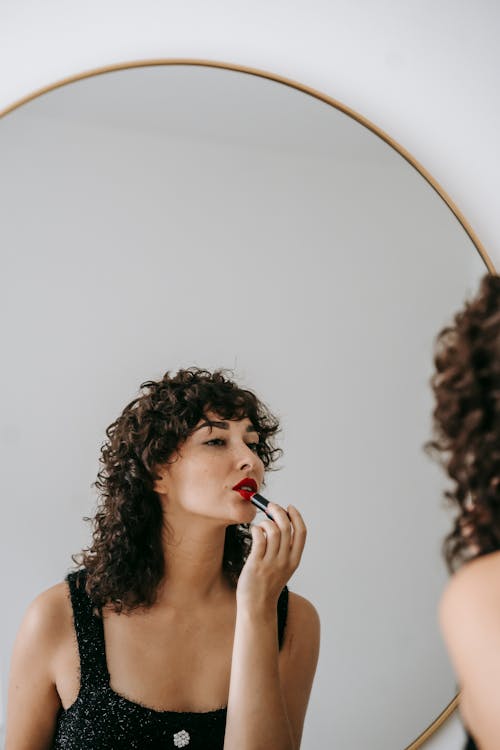 Charming female with dark curly hair wearing black outfit rouging lips while looking at mirror in room during beauty routine