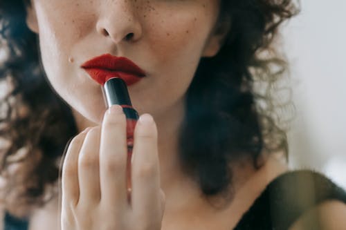 Unrecognizable elegant female with dark curly hair and freckles applying bright makeup on lips in room while preparing for event