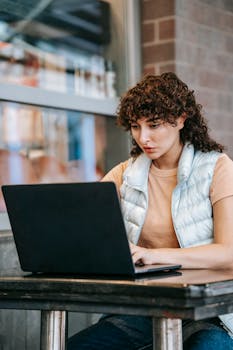 Adult attentive female remote employee typing on netbook while working on project at cafe table
