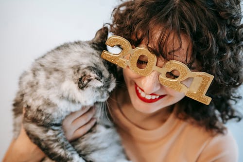 Crop candid female in decorative glasses with numbers embracing fluffy cat with closed eyes during festive event on white background