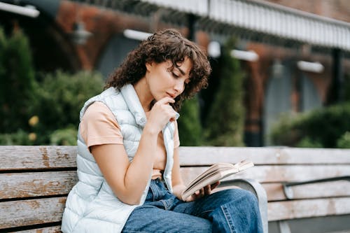 Free Adult thoughtful female touching lips while reading textbook and sitting with crossed legs on wooden bench in town Stock Photo