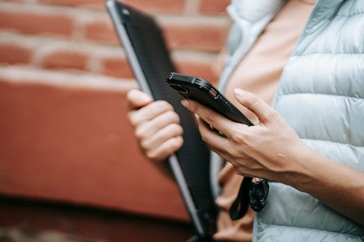 Close-up of a woman using a smartphone and holding a laptop against a brick wall.