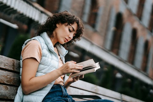 Free Young concentrated female in casual outfit sitting on bench on old city street and reading interesting novel Stock Photo