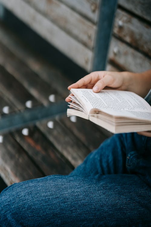 Free From above of crop unrecognizable person in casual jeans sitting on wooden bench and reading book in daytime Stock Photo