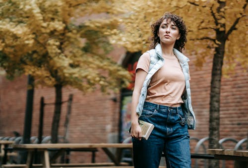 Free Pensive female in casual clothes with book in hand walking on city street against brick building and trees with autumn foliage in daytime Stock Photo