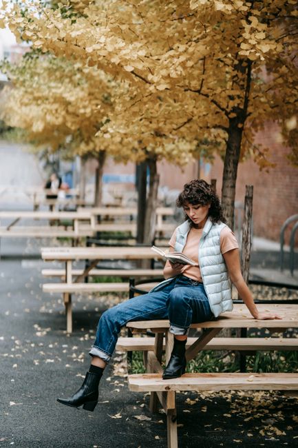 Pensive woman in trendy clothes sitting on wooden table and reading book on city street with autumn trees in daytime