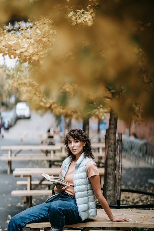 Free Stylish woman with short curly hair sitting on wooden table on street among trees with autumn foliage and looking at camera while reading book in daytime Stock Photo