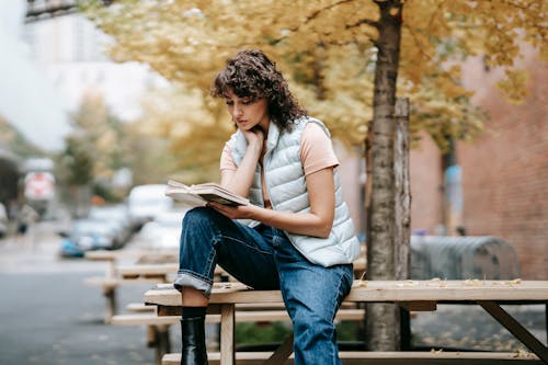 Pensive female in trendy outfit reading book while sitting on wooden table near autumn tree on city street in daytime