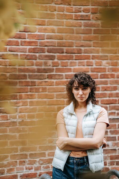 Free Confident female with curly hair in casual outfit standing with crossed arms on street against brick wall and looking at camera in daytime Stock Photo