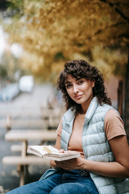 Free Smiling curly haired female in casual clothes sitting on bench while reading book and looking at camera Stock Photo
