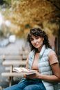 Smiling curly haired female in casual clothes sitting on bench while reading book and looking at camera