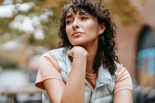 Low angle of crop pensive female with dark curly hair touching chin while looking away