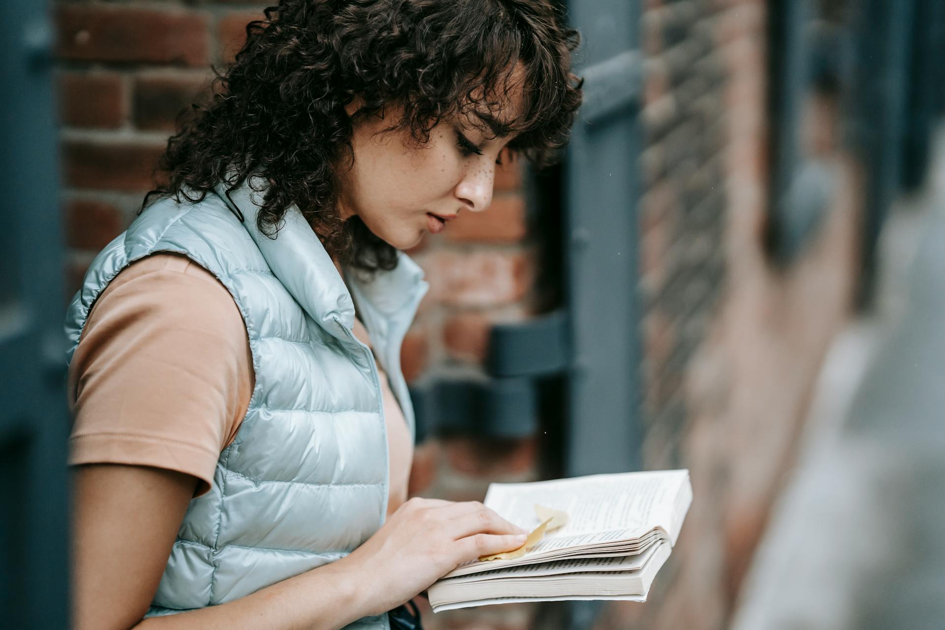 Side view of crop concentrated female standing near brick building and reading interesting story in book while spending time in city