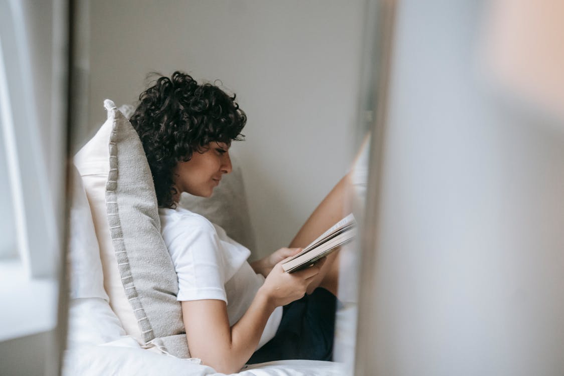 Free Side view of young curly haired female in sleepwear relaxing on soft bed with pillows while reading paper book Stock Photo
