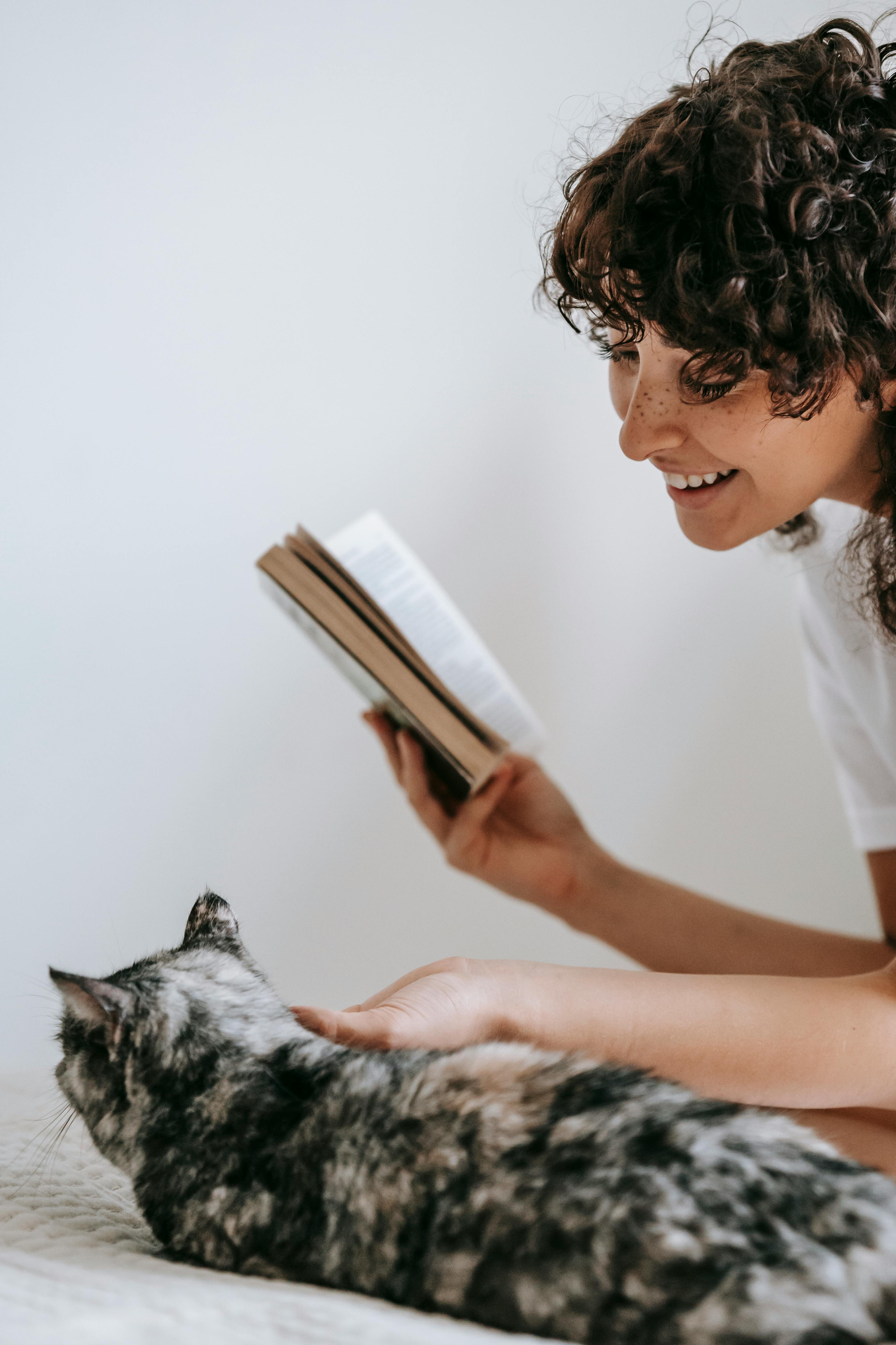 Side view of crop positive female reading book and caressing fluffy cat lying on bed
