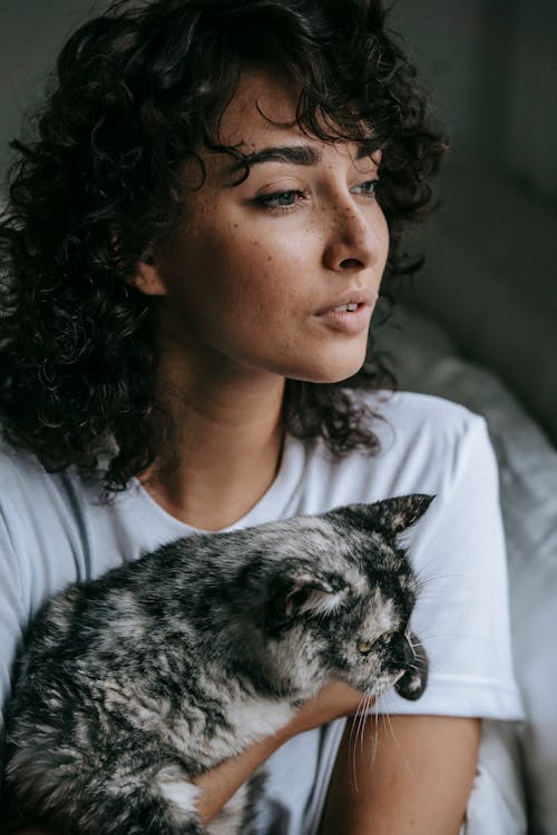 Crop dreamy young female with long curly hair and freckles sitting on bed with cat and looking away