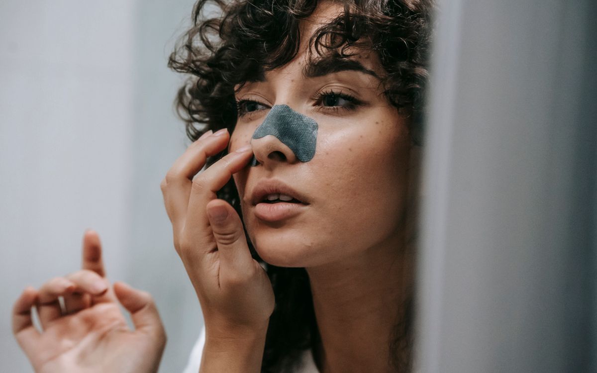 Mirror reflection of young female with curly hair putting nose strip mask on face in bathroom