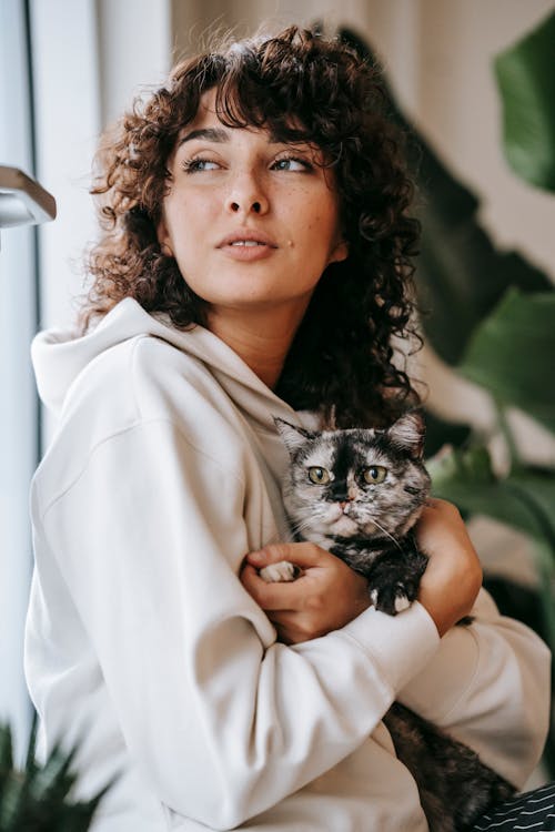 Attractive female owner in white sweater looking away while sitting with dappled cat in flat with green plants on blurred background