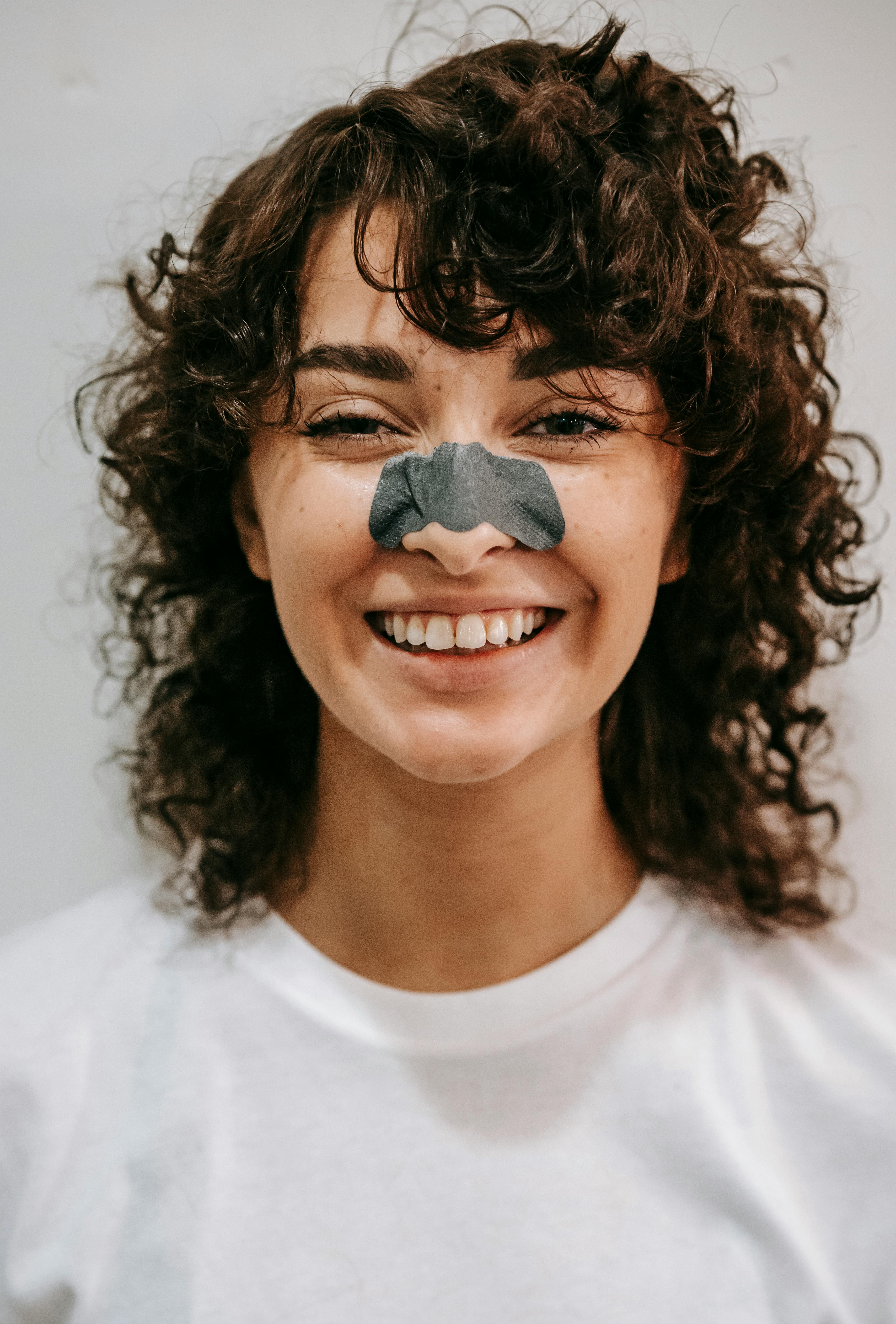 Cheerful female with curly hair and blackhead cleanse nose strip wearing white t shirt looking at camera on gray background