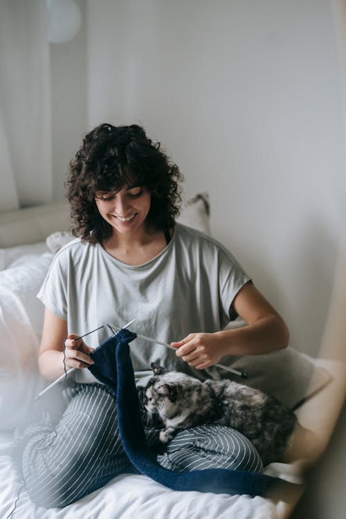 Free Full body of positive female owner with dappled cat sitting on bed with knitting needles and blue thread in bedroom Stock Photo