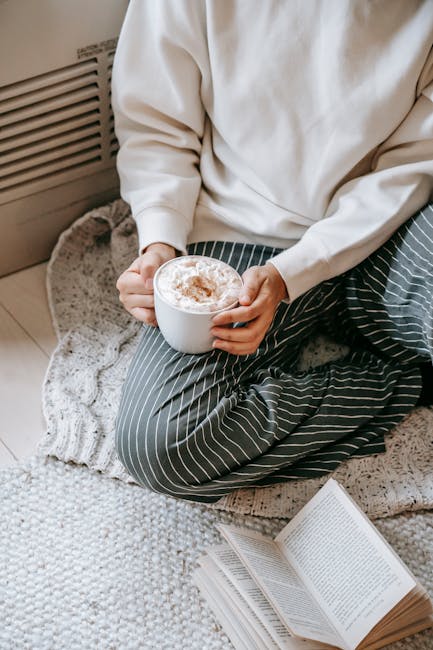 From above of unrecognizable female sitting on plaid with cocoa topped with marshmallow near opened book during weekend in cozy apartment
