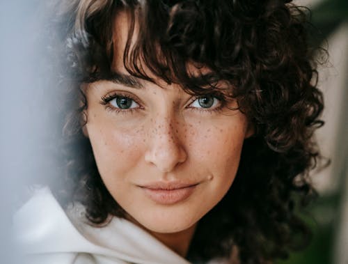 Headshot of attractive female with dark curly hair looking at camera against green leaves on blurred background in light room