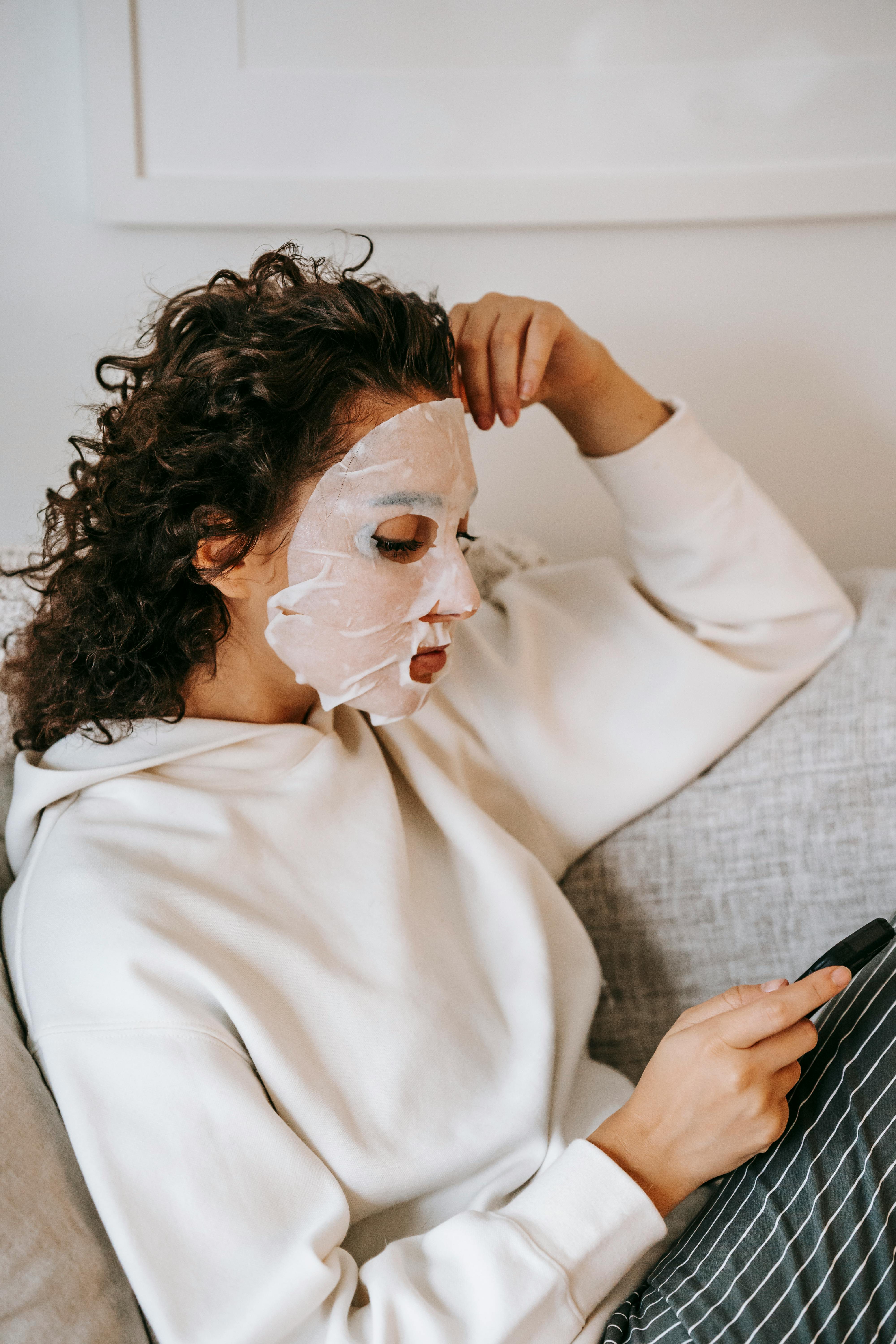 pensive woman in sheet mask browsing smartphone