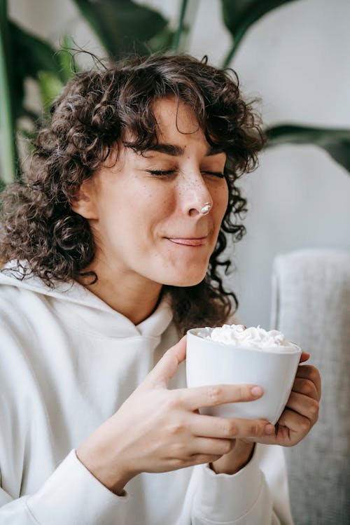 Delighted female with curly hair and closed eyes licking lips while drinking hot beverage topped with marshmallow in room against blurred background