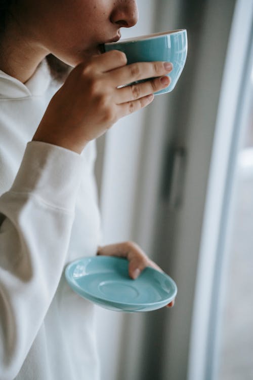 Free Unrecognizable peaceful female drinking hot beverage from ceramic mug while standing near window in light room on blurred background during weekend Stock Photo