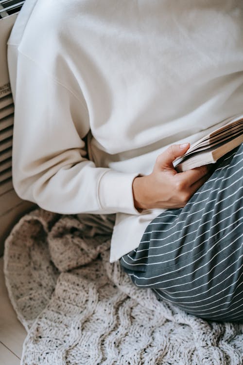 From above of crop anonymous female reading interesting story in novel while resting on knitted woolen plaid