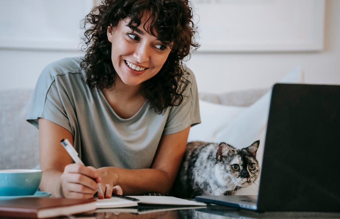 Young female smiling and taking notes in organizer while cat watching on netbook at table at home