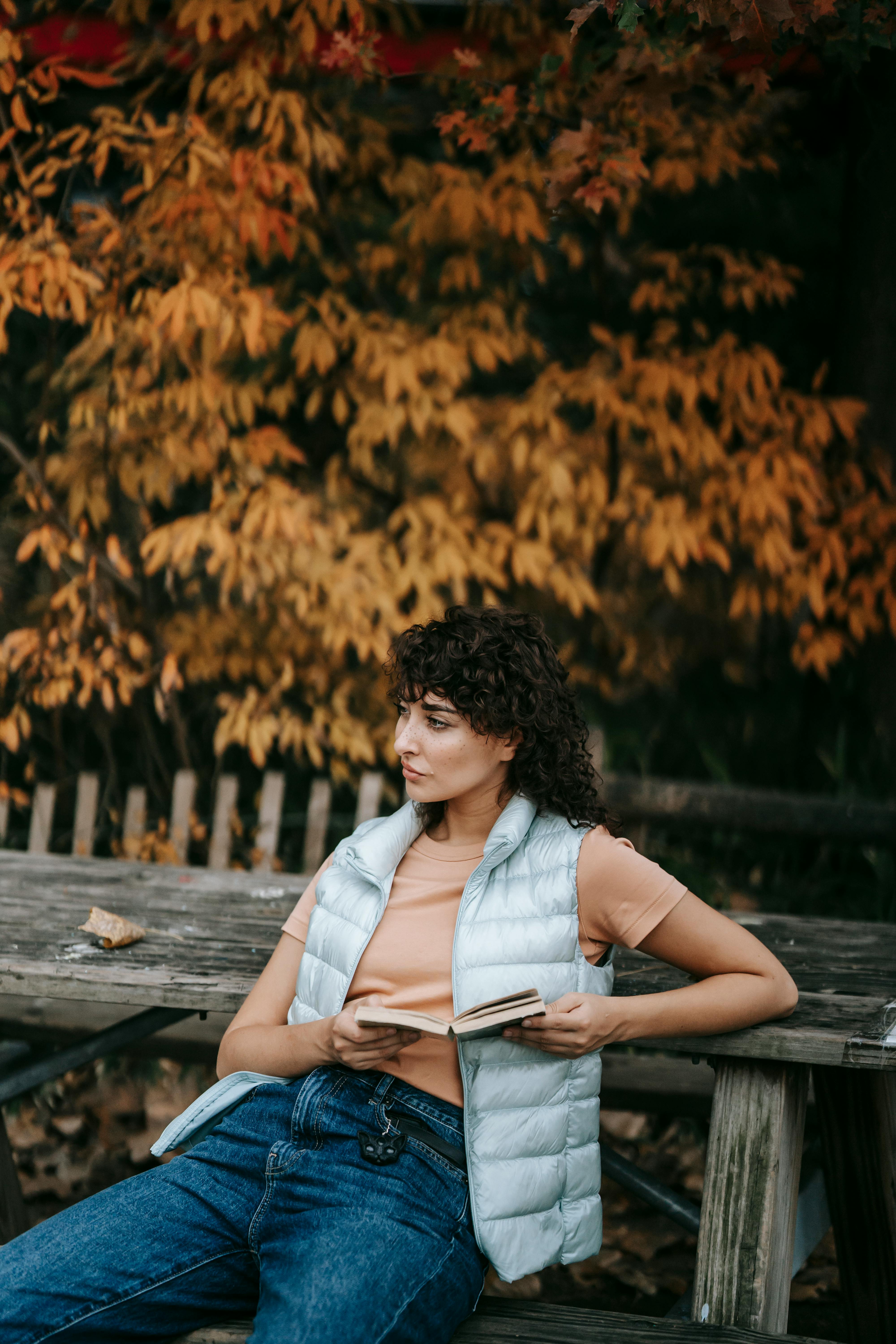 Pensive woman with dark curly hair in casual clothes sitting with book in hands on wooden bench against autumn trees in park and looking away in daytime