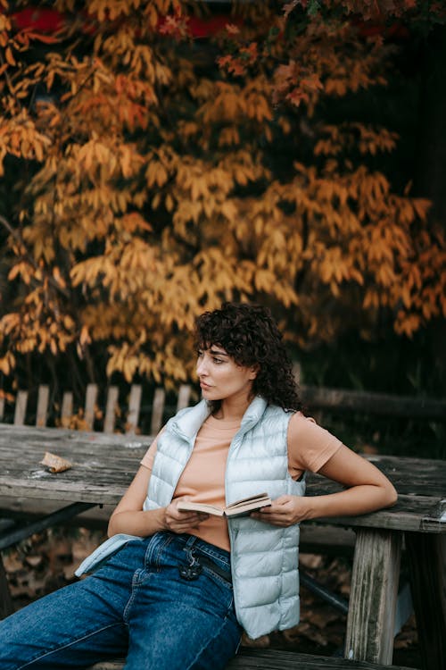 Pensive woman with dark curly hair in casual clothes sitting with book in hands on wooden bench against autumn trees in park and looking away in daytime