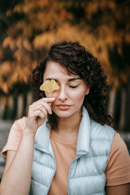 Charming woman with curly hair in casual clothes covering eye with yellow leaf against autumn tree in park in daylight
