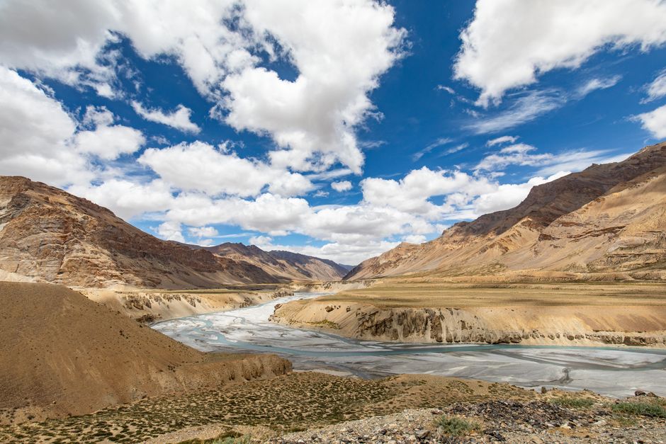 Expansive view of Ladakh's rugged terrain and Indus River under a vibrant blue sky with white clouds.