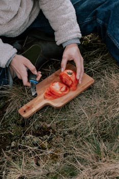 Person slicing tomatoes on wooden board in natural outdoor setting. Perfect for food preparation, picnic, or outdoor cooking themes.