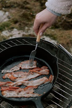 Close-up of bacon strips frying in a pan on a charcoal grill outdoors.