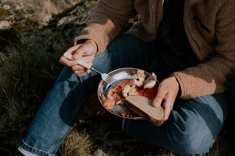 A Person In Brown Coat Sitting On The Ground While Eating