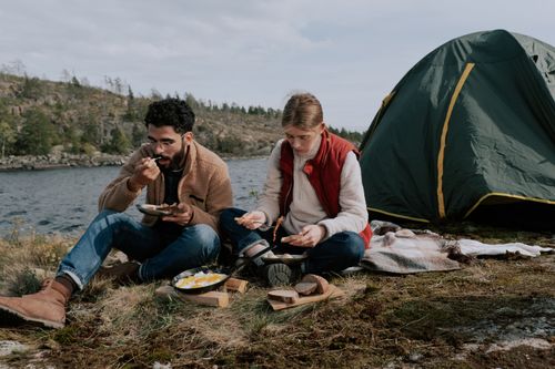 Free A couple sits by their tent enjoying a meal by a lakeside campsite during a day in nature. Stock Photo