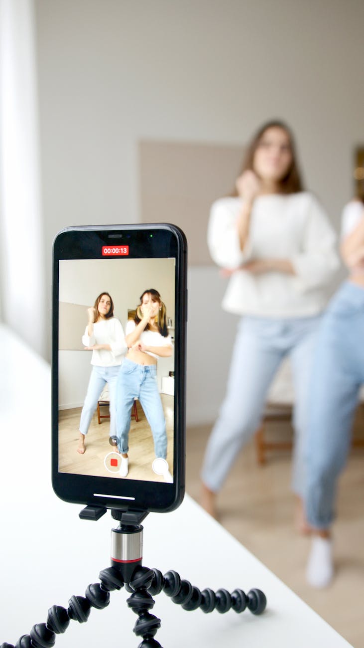 Two young women dancing in front of a smartphone camera on a tripod indoors.